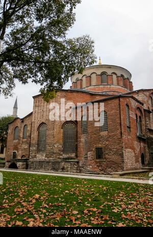 Vista esterna e da un basso angolo di abside sul lato est della pietra e mattoni costruito Aya Irini (Hagia Irene), con un minareto sullo sfondo a sinistra e lascia su erba verde e un marciapiede in primo piano, a Aya Irini, un ex greco chiesa ortodossa orientale, ora adibito a museo e sala concerti, situato nel cortile esterno del palazzo Topkapi a Istanbul, Turchia, 16 novembre 2017. () Foto Stock