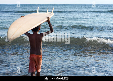 Un locale giovane ragazzo indonesiano in pantaloncini usurato porta il suo donato tavole da surf sulla sua testa in un mare calmo.