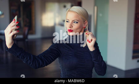 Elegante giovane donna bionda tenendo selfie a casa in ambienti interni Foto Stock