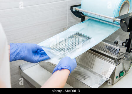 Chiudere l assistente in mani guanti imballaggio strumento dentale impostato per il trattamento in autoclave in un sacchetto di plastica utilizzando il vuoto macchina impacchettatrice. Sterilità e sicurezza il concetto di cura. Messa a fuoco selettiva. Foto Stock