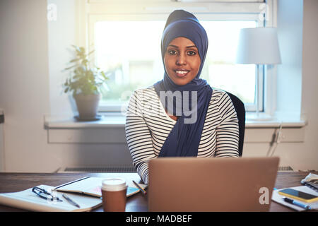 Sorridente giovane musulmano imprenditore femmina indossando un hijab seduto nel suo ufficio a casa a lavorare online con un computer portatile Foto Stock