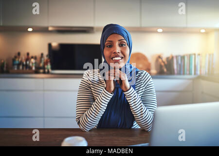 Certi giovani arabi imprenditore femmina indossando un hijab lavorando su un computer portatile mentre si siede a un tavolo nella sua cucina Foto Stock