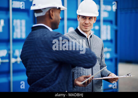 Sorridente freight manager indossando un hardhat in piedi da contenitori di trasporto merci su una grande spedizione commerciale dock parlando con un collega Foto Stock