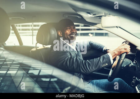 Imprenditore africani che indossa un blazer e sorridere mentre alla guida della sua auto durante i suoi spostamenti di mattina attraverso la città Foto Stock