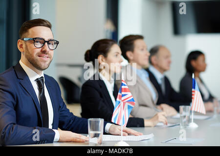 Elegante delegato in tuta e occhiali guardando la fotocamera alla conferenza politica sullo sfondo di altri partecipanti Foto Stock