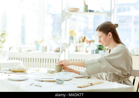 Giovane donna seduta al tavolo e fare argilla o tazza di ceramica nel suo lavoro in studio Foto Stock