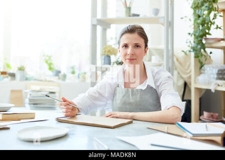 I giovani creativi donna in maglia bianca e grembiule grigio seduti a tavola e delineando nel Blocco note mentre il brainstorming Foto Stock