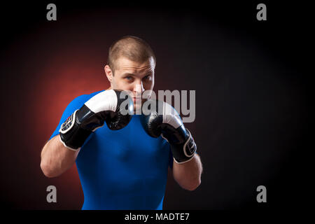 Un forte dai capelli scuri boxer sportman in un blu di indossare abbigliamento sportivo rush guard e in bianco e nero i Guantoni boxe contro un rosso a luci su un nero i Foto Stock