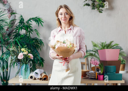 Ritratto di fiorista sorridente nel grembiule con bouquet di fiori con carta kraft su sfondo di piante da interni in camera Foto Stock