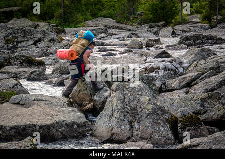 Uomo attraversa un fiume di montagna con un grande zaino pesante sulle rocce. Foto Stock