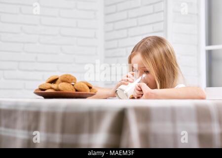 Carino bambina con dritto bionda hairis bere latte da vetro trasparente vicino al piatto pieno di dolce, deliziosi biscotti. La tabella è coperto con plaid tovaglia marrone. Foto Stock