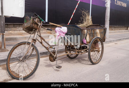 Pechino, Cina - 26 Aprile 2010: primo piano della spazzatrice marrone triciclo in metallo con la sua scopa, parcheggiate lungo la strada. Nero e viola parete. Foto Stock