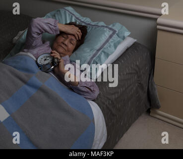 Donna senior con dolorose mal di testa tenendo la sua sveglia durante le ore notturne Foto Stock