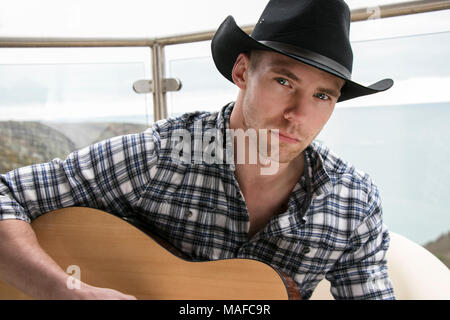 Bel paese e cowboy western con la cantante che indossa un cappello e a suonare la chitarra su hotel balcone con vista oceano in background Foto Stock