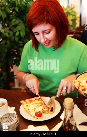 Giovane donna di mangiare la pizza in cafe. Ella taglia una fetta di pizza con un coltello. Foto Stock