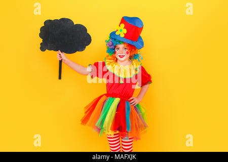 La felicità ragazza in costume da clown e hat holding tavola nera. Studio shot, isolato su sfondo giallo Foto Stock