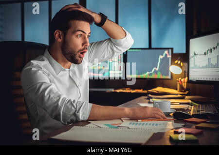 Giovane uomo barbuto trader in ufficio seduta a tavola che stanno guardando sullo schermo toccando i capelli scioccato Foto Stock