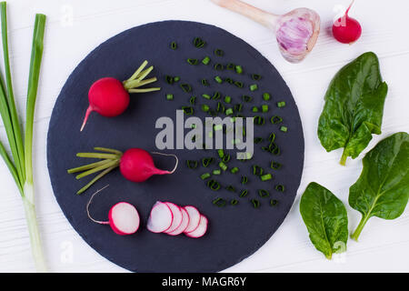 Cucina in legno tavolo con sani ingredienti cottura. Tondo nero ardesia e il cipollotto, basilico, aglio e ravanelli in legno bianco. Molla di cottura sal Foto Stock