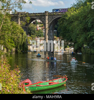 Persone e rilassanti gite in barca a remi sul fiume Nidd sotto il cielo estivo blu, come il treno passa sul ponte in scenic sunny Knaresborough, England, Regno Unito Foto Stock