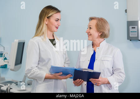 Senior medico parlando con la giovane donna in piedi assistente in ufficio ginecologiche con sedia e lampada sullo sfondo Foto Stock