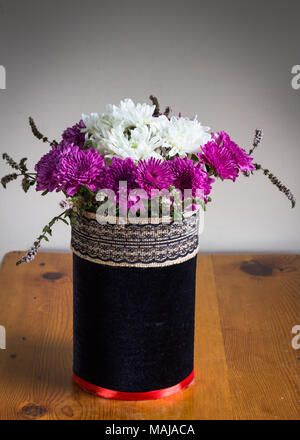 Chrysanthenums in un rustico di velluto nero vaso coperto con nastro ricamato band - viola e bianco floreale sul tavolo di legno Foto Stock