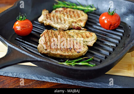 Le bistecche grigliate in una padella di ferro con pomodori e il ramo di rosmarino. Bistecche grigliate su un tavolo in legno, cucina gourmet. Foto Stock