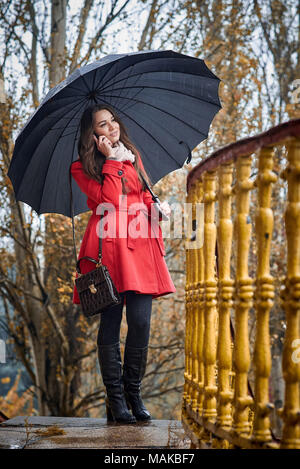 Una ragazza in un cappotto rosso sotto un ombrello nero sta parlando al telefono Foto Stock