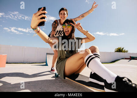 La donna a cavallo su skateboard tenendo selfie con amici spingendo da dietro. Amici di sesso femminile facendo una selfie mentre cavalcate skateboard a skate park. Foto Stock