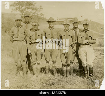 Orizzontale, seppia fotografia che mostra sette uomini in uniforme in piedi in una linea che posano per una foto di gruppo. Gli uomini sono in piedi in mezzo a un campo di erba e gli edifici sono visibili in lontananza. Titolo: sette soldati in uniforme in posa di una linea per una foto di gruppo. . Tra circa 1914 e circa 1918. Michel, Carl Foto Stock