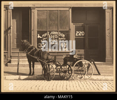 In orizzontale e di fotografia in bianco e nero di un a cavallo il buggy parcheggiato contro un marciapiede davanti a un negozio di fronte che legge 'Mike Johnson Stivali e scarpe". Titolo: Mike Johnson Shoe Repair. . Tra circa 1800 e circa 1900. Swekosky, William G., 1895-1964 Foto Stock