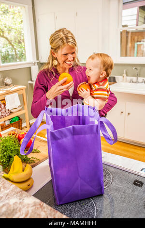 Una madre con i suoi dieci mesi di old boy nella loro cucina domestica di disimballare i generi alimentari. Foto Stock