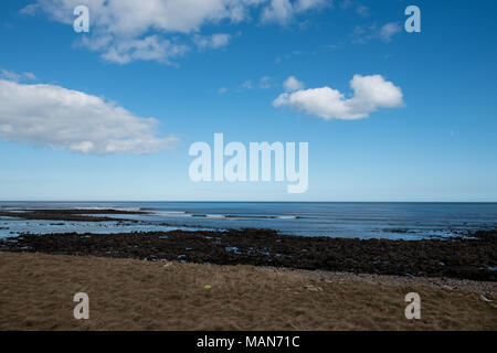 Costa rocciosa con bassa marea sotto un cielo blu brillante con nuvole sparse sulla costa del Regno Unito, rivelando pozze di marea e texture costiere Foto Stock