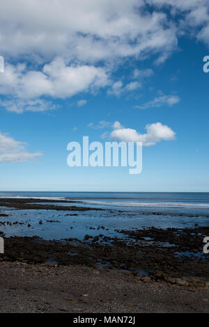 Costa rocciosa con bassa marea sotto un cielo blu brillante con nuvole sparse sulla costa del Regno Unito, rivelando pozze di marea e texture costiere Foto Stock