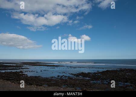 Costa rocciosa con bassa marea sotto un cielo blu brillante con nuvole sparse sulla costa del Regno Unito, rivelando pozze di marea e texture costiere Foto Stock