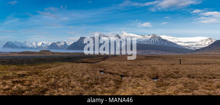 Panorama di montagne innevate vicino Skalafell in Islanda con cielo blu, erba, un flusso e una strada tortuosa in primo piano Foto Stock