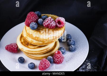 Pila di pancake con lamponi e mirtilli rivestite con zucchero a velo sulla piastra bianca e sfondo nero Foto Stock