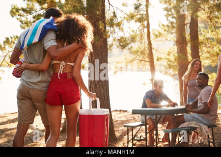 Giovane con una scatola fredda a piedi amici parlando a tavola accanto al lago Foto Stock