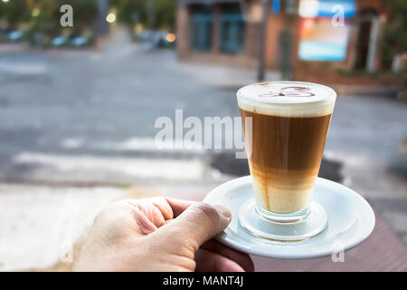 Un maschio di mano in mano un bicchiere con cappuccino caldo su una piastra bianca con la chiave di violino sulla parte superiore, sfondo all'aperto. Foto Stock