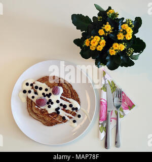 Una piastra di Pancake fatti in casa serviti con yogurt e frutti di bosco. Vi è anche vasellame e bellissimo fiore giallo nell'immagine. Foto Stock