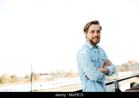 Casual bella bionda uomo in piedi fuori in maglietta blu, sorridente e avere un atteggiamento positivo, cercando di fotocamera con bracci ripiegati Foto Stock