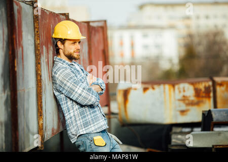 Uomo forte con la barba in plaid shirt, sito lavoratore con il casco in testa, con strumenti, appoggiarsi contro alcuni oggetti arrugginiti, attesa per la fine del giorno, wi Foto Stock