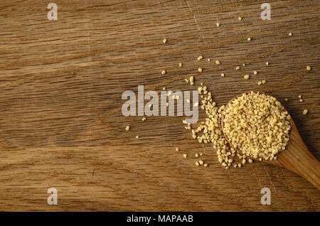 Scatto dall'alto di un cucchiaio di legno, riempito con couscous crudo grani, versarsi alla vecchia, graffiato la superficie in legno di quercia. Foto Stock