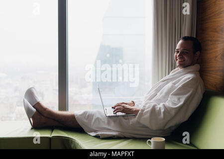 Giovane imprenditore sorridente lavorando sul computer portatile bianco da indossare accappatoio seduta vicino la finestra con tazza di caffè guardando sunrise vista città. Concetto di motivazione Foto Stock