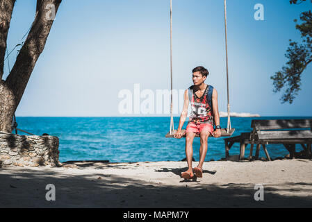 Giovane uomo nel serbatoio superiore si siede su una corda swing sulla riva sabbiosa contro il blu del mare e cielo chiaro. Ko Samet (Koh Samed), Tailandia Foto Stock