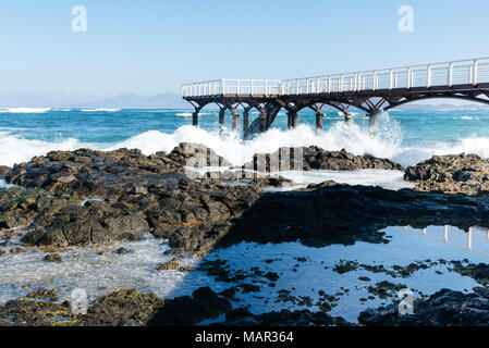 Onde che si infrangono contro molo sulla spiaggia di Corralejo al mattino contro il cielo blu Foto Stock