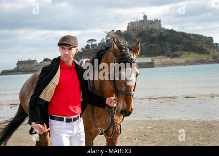 Bel cavallo maschio conducente in piedi camminando il suo cavallo, indossando il tappo piatto, pantaloni bianchi e stivali neri sulla spiaggia di fronte al St Michael's Mount Foto Stock
