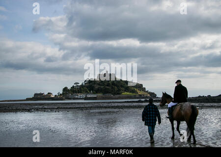 Cavallo cavaliere a cavallo attraverso la spiaggia di St Michael's Mount con trainer camminando lungo il lato. Foto Stock