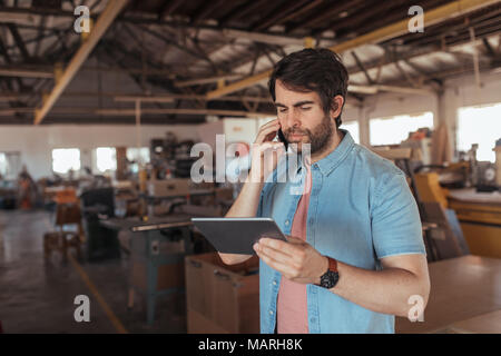 Falegname utilizzando un telefono e tablet nella sua officina di grandi dimensioni Foto Stock