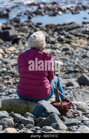 Un vecchio o invecchiamento anziana signora seduta su di una spiaggia profonda nel pensiero e pensando che guarda al mare attraverso le rocce e piscine di roccia. Grigio donna dai capelli. Foto Stock