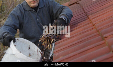 Uomo di pulizia grondaia sporca da muschi e foglie. Edificio con impuro tetto di tegole dopo l'inverno. Pulizia di Primavera. Foto Stock